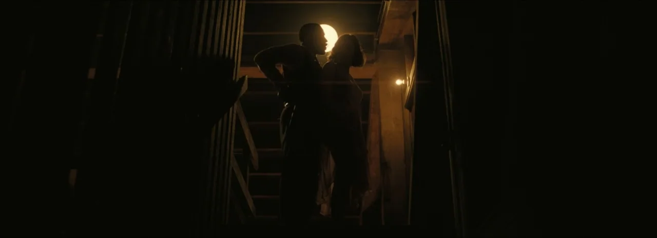 Sinners (2025) movie still: fullbody — A dimly lit wooden staircase features a man and woman standing near a lamp. The couple is positioned against…