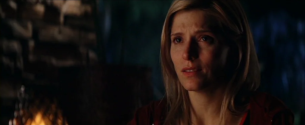 Magnolia (1999) movie still: closeup — A dark indoor setting features a stone wall illuminated by a low-level orange flame. A woman with blonde hair…