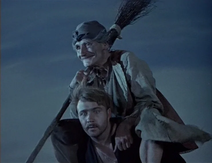 Viy (1967) movie still: medium — A featureless dark blue sky serves as the setting for the two figures. A young man carries an elderly woman on his…