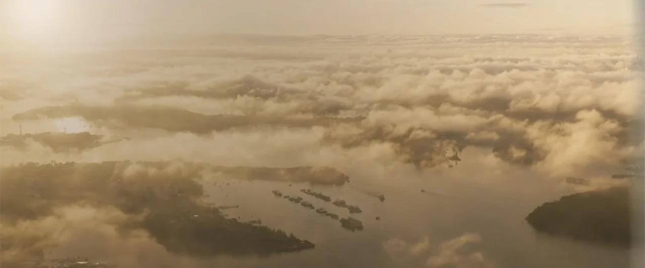 Unsung Hero (2024) movie still: establishing — A wide aerial view of a calm harbor obscured by a thick blanket of fog. Small boats are visible…
