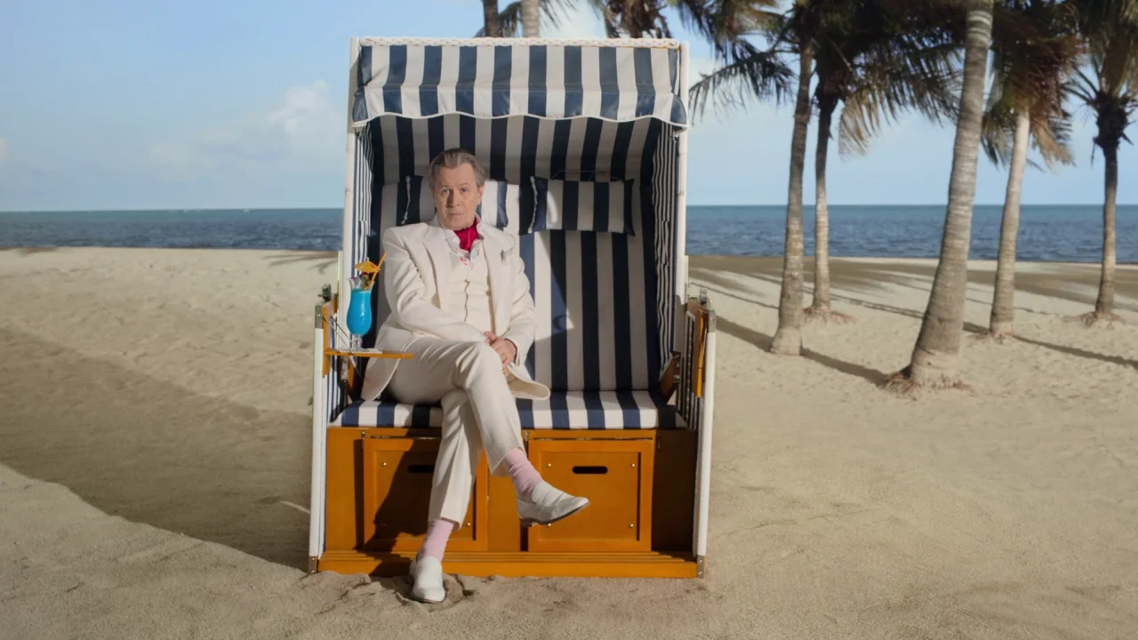 The Laundromat (2019) movie still: fullbody — A man sits in a blue and white striped beach chair on sand. Tall palm trees stand in the background…