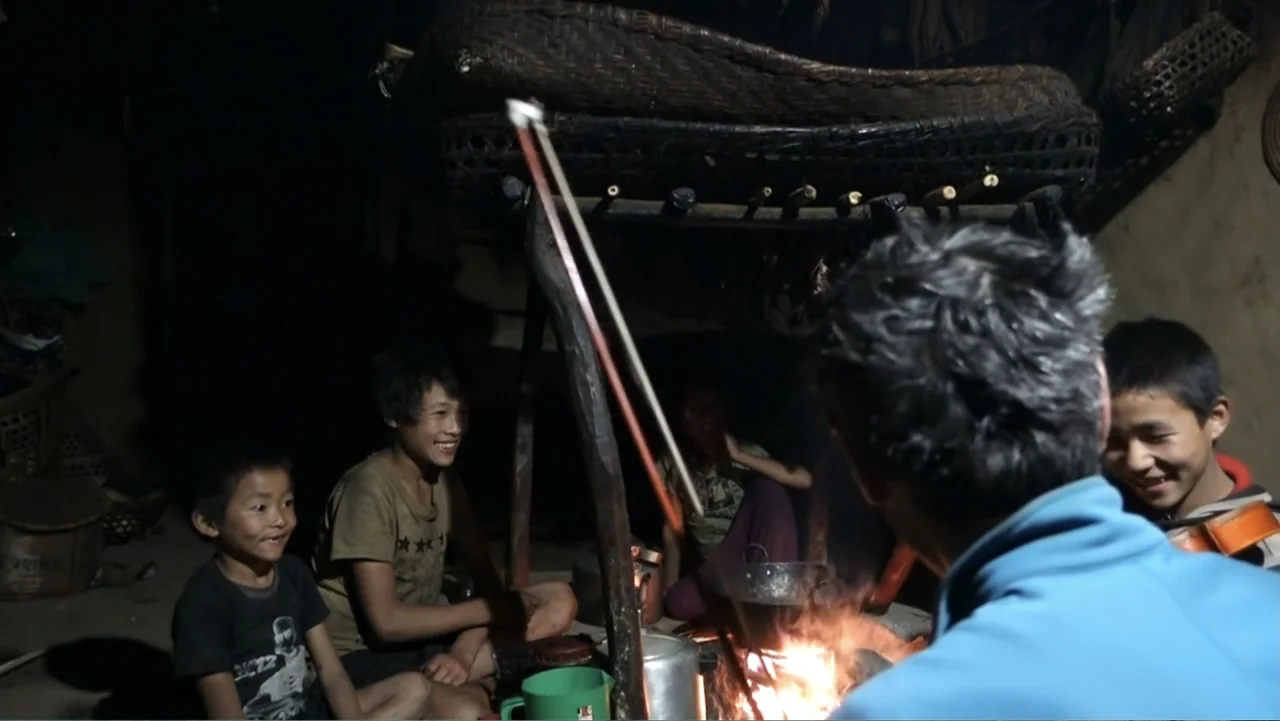 Hondar 2050 (2018) movie still: medium — A dark, rustic interior features a central fire pit beneath a suspended drying rack. A man plays violin…