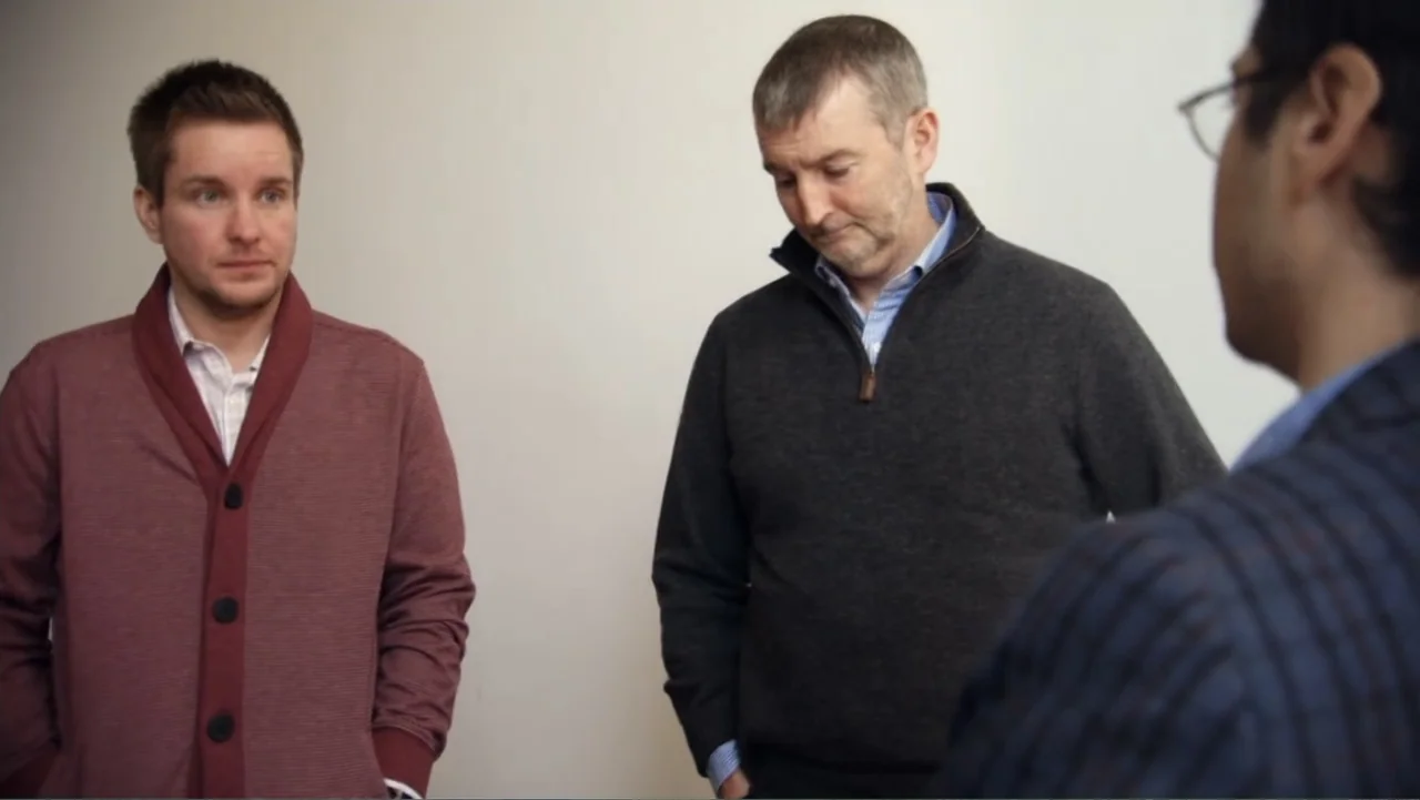 Suited (2016) movie still: medium — An interior space with plain white walls serves as the backdrop for the meeting. Three men stand together in a…