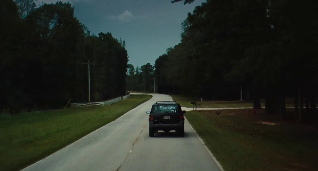 The Giant (2019) movie still: wide — A rural paved road winds through a dense corridor of trees under overcast skies. A dark SUV with a painted…