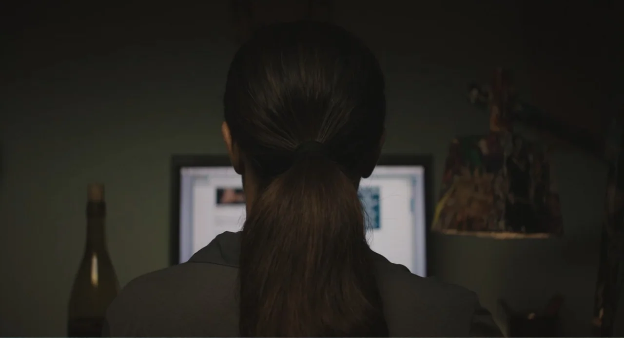 Beatriz at Dinner (2017) movie still: medium — A dimly lit room features a person viewed from behind looking at a monitor. The screen glows brightly…