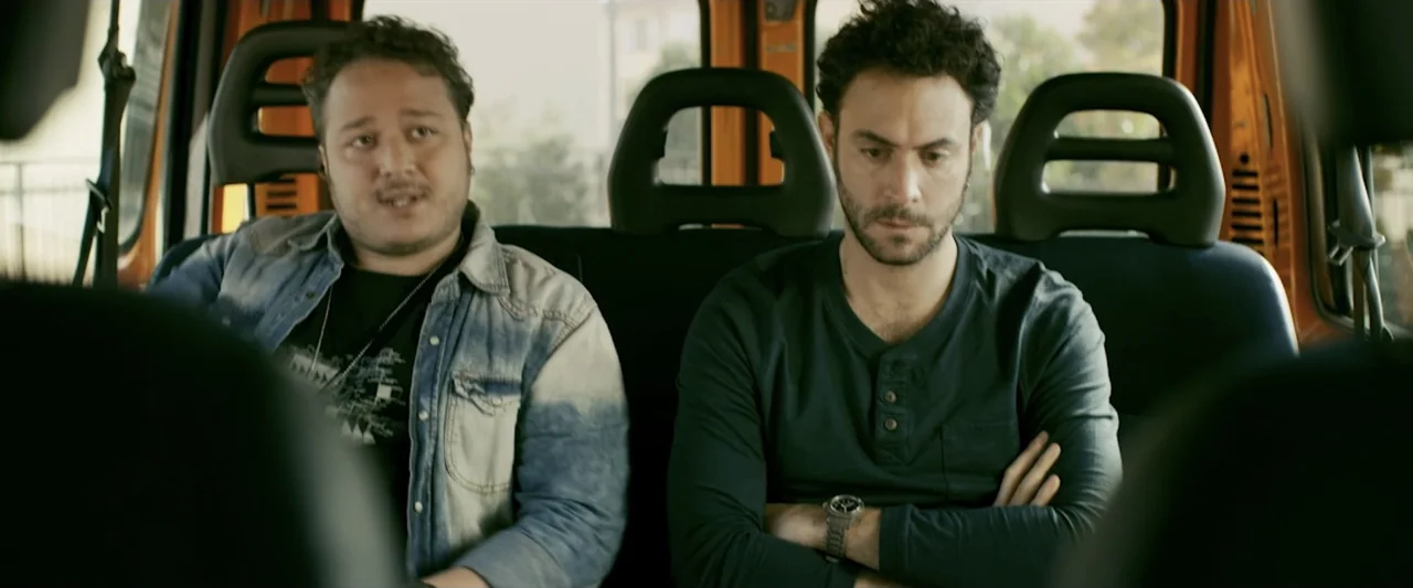 Taranta On the Road (2017) movie still: medium — The back seats of an orange van provide a cramped and utilitarian setting. Two men sit side by side…