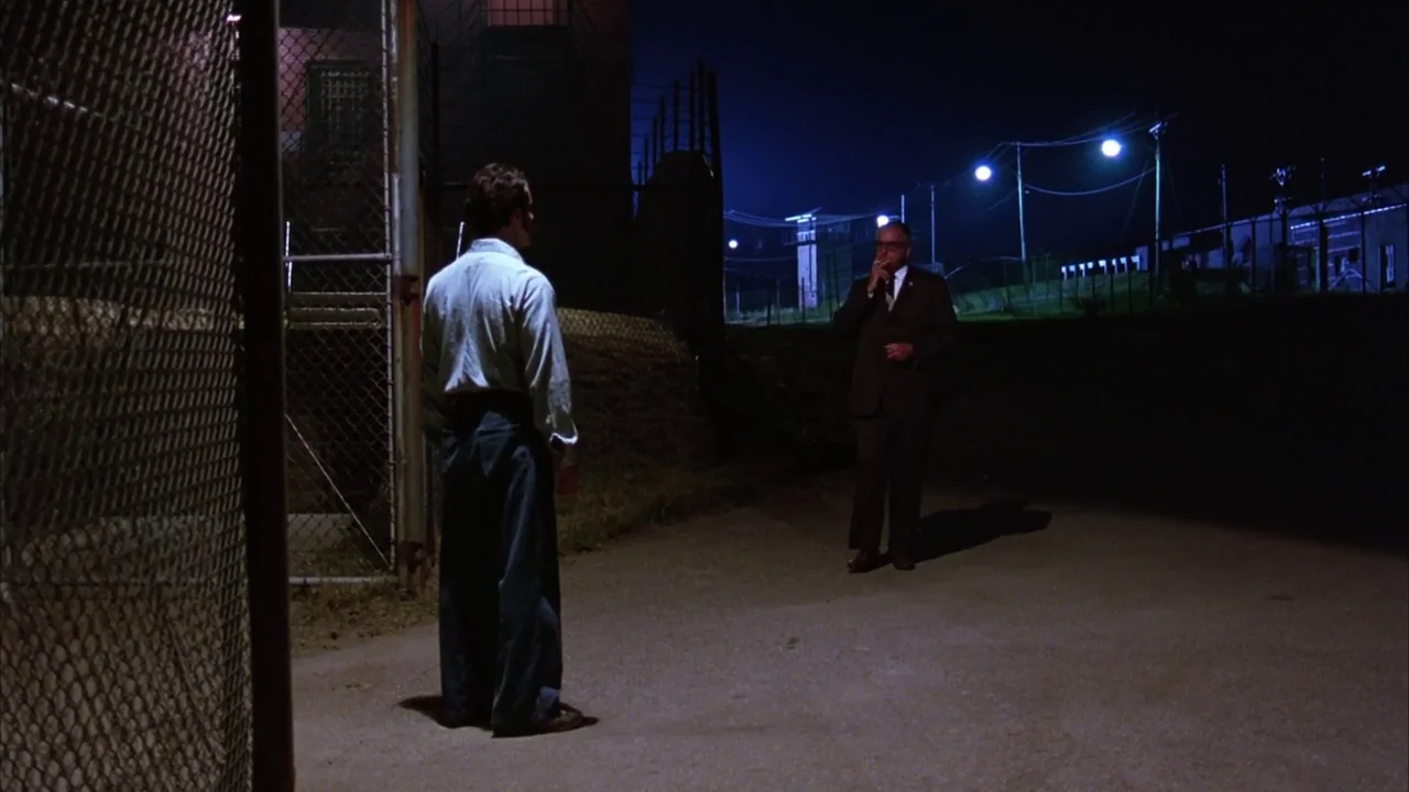 The Shawshank Redemption (1994) movie still: wide — A night-time view of a high-security enclosure illuminated by bright overhead industrial street…
