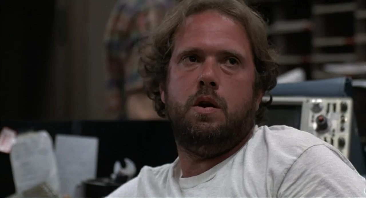 WarGames (1983) movie still: closeup — An interior office space with blurred technical equipment and filing structures in the background. A man with…