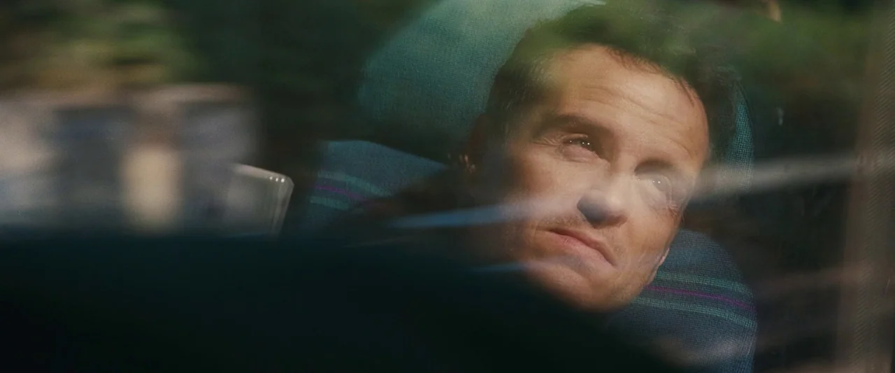 All Of Us Strangers (2023) movie still: closeup — A man sits in a train seat while viewed through a reflective window. The blue patterned fabric of…