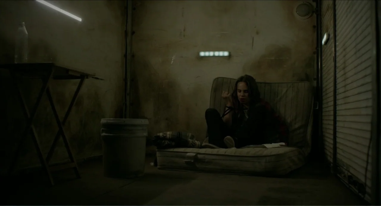 Instantes Capturados (2022) movie still: wide — A dimly lit concrete room features a mattress on the floor and industrial walls. A young woman with…