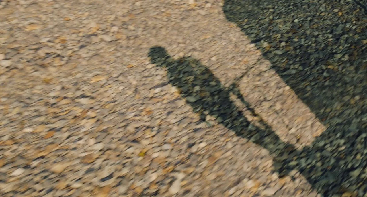 Train To Busan movie still: wide — A gravel path surface is captured from above during a sunny day. A person's dark shadow moves across the textured…