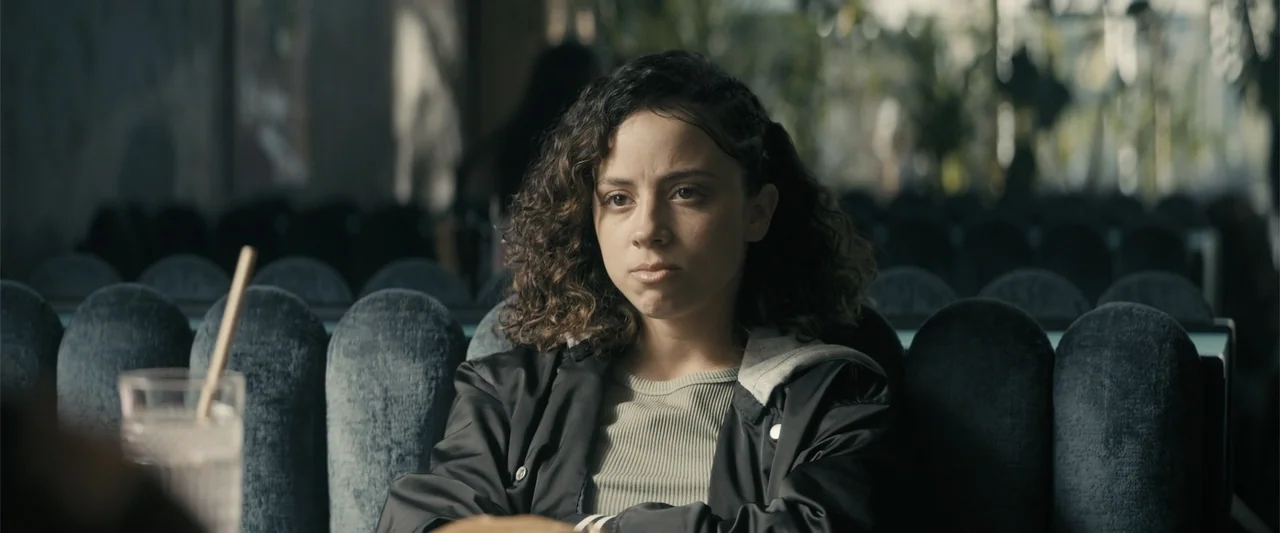 Perfect Addiction (2023) movie still: medium — A modern booth inside a restaurant has blue tufted velvet seats. A young woman with curly hair sits…