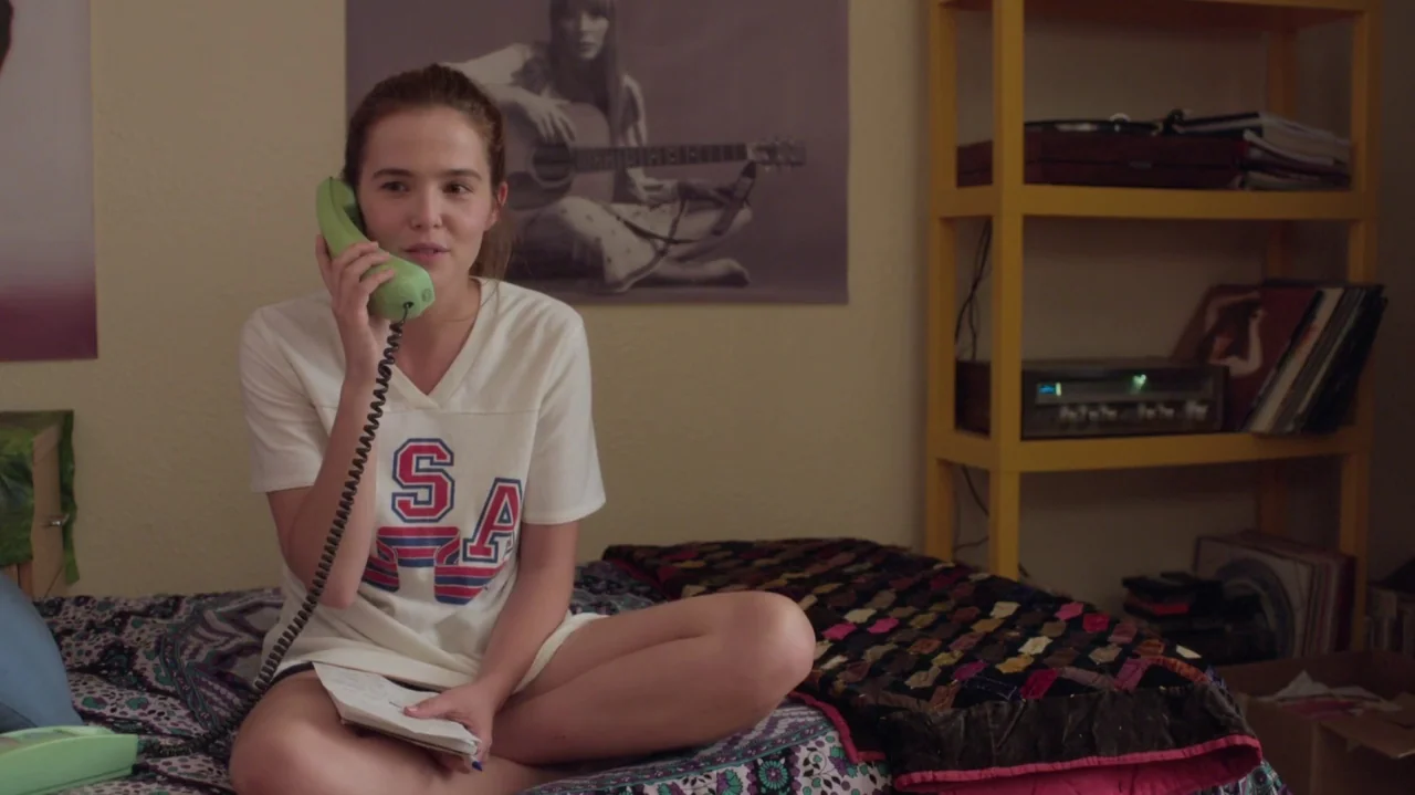 Everybody Wants Some!! (2016) movie still: medium — A teenage girl sits cross-legged on a bed in a room decorated with posters. She speaks into a…