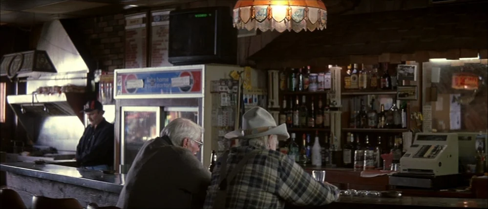 The Straight Story (1999) movie still: medium — A dimly lit rural tavern with wood-paneled walls and a liquor-stocked back bar. Two elderly men sit…