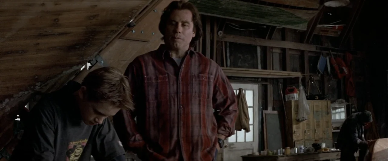 Angst (2017) movie still: medium — A cluttered wooden workshop has exposed roof beams and various tools stored in lockers. A man watches over a boy…