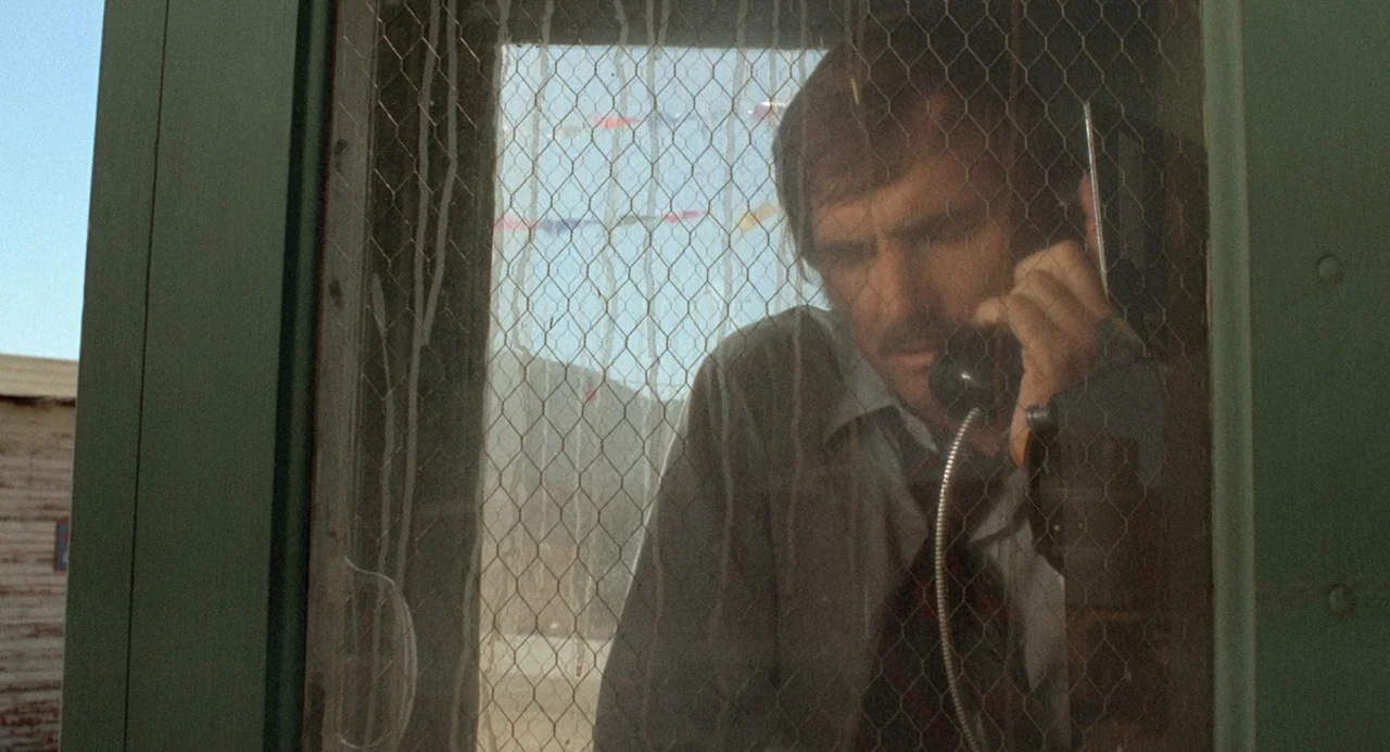 Duel (1971) movie still: medium — A man stands inside a vintage outdoor glass-paneled phone booth. He holds the telephone receiver to his ear while…