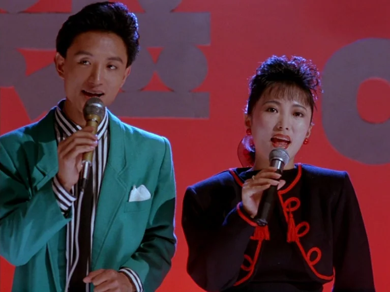 Black Snow (1990) movie still: medium — A man and a woman perform on a stage with a bold red background. They both hold microphones while singing…
