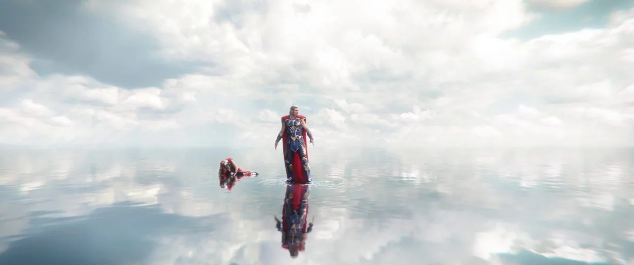 Thor: Love and Thunder (2022) movie still: wide — An expanse of calm, reflective water mirrors a bright sky filled with heavy clouds. A costumed man…