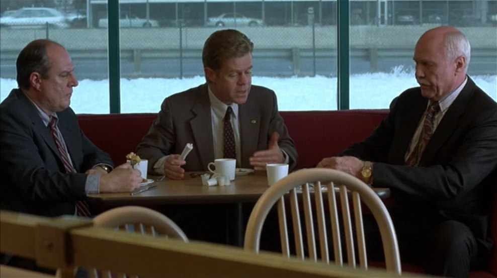 Fargo (1996) movie still: medium — A diner booth with a red vinyl seat sits in front of large windows. Three men in professional business attire sit…