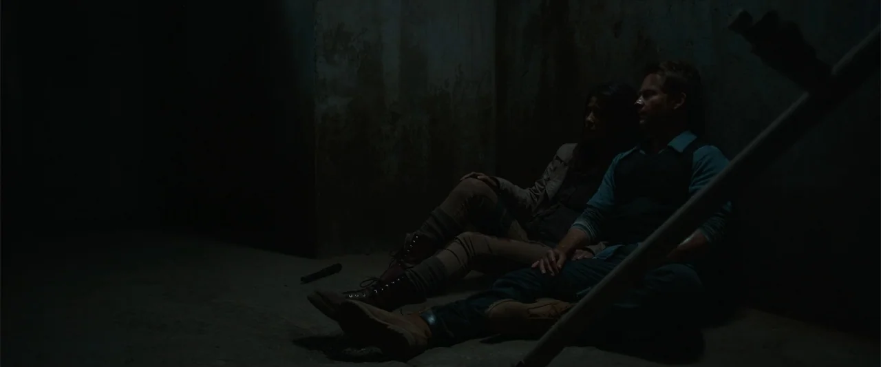 The Condemned 2 (2015) movie still: fullbody — A dark concrete enclosure with stained walls and a barren floor. Two people sit slumped against the…