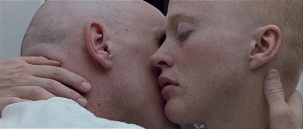 THX 1138 (1971) movie still: closeup — Two individuals are positioned close together in a neutral interior space. A man and a woman are kissing with…