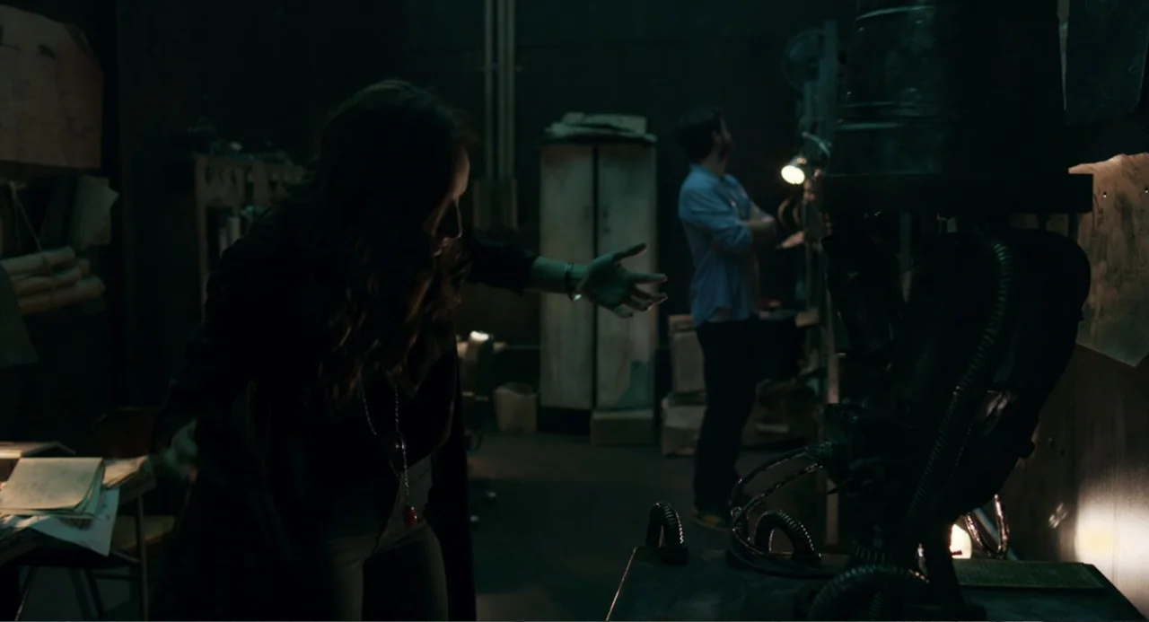 The Lift (2021) movie still: medium — A cluttered, dimly lit industrial space contains various technical tools and machinery components. A woman…
