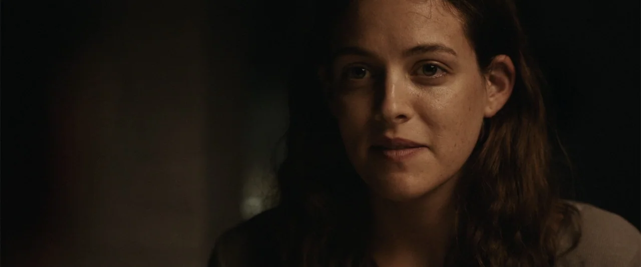 It Comes at Night (2017) movie still: closeup — A dark indoor space with minimal detail visible in the background shadow. A young woman with brown…