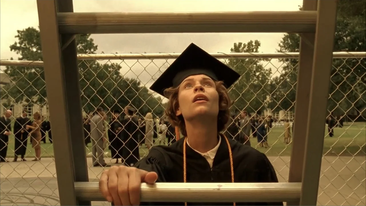 Temple Grandin (2010) movie still: medium — A student in academic regalia stands behind a chain-link fence on a grassy campus. The graduate looks…