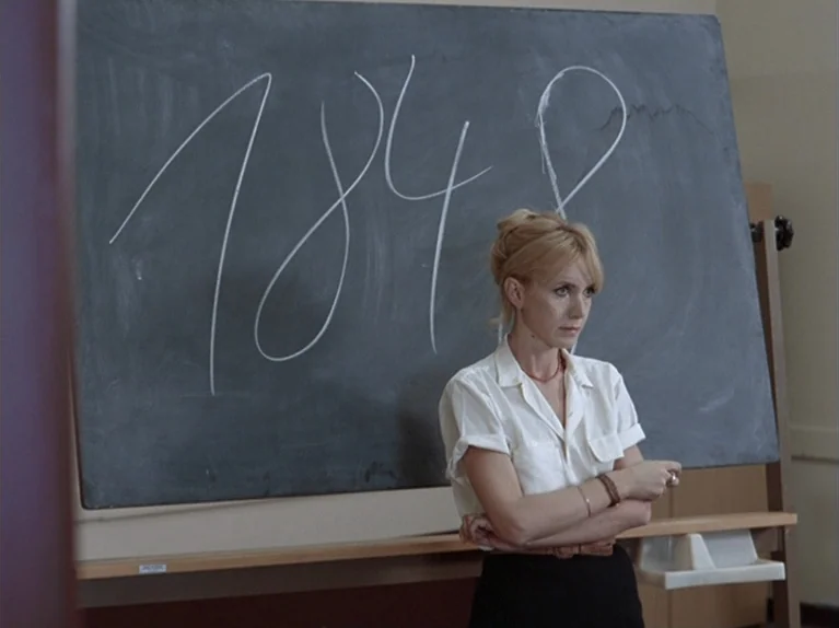 The Third Generation (1979) movie still: medium — A woman stands before a large black slate board featuring white handwritten numbers. She wears a…
