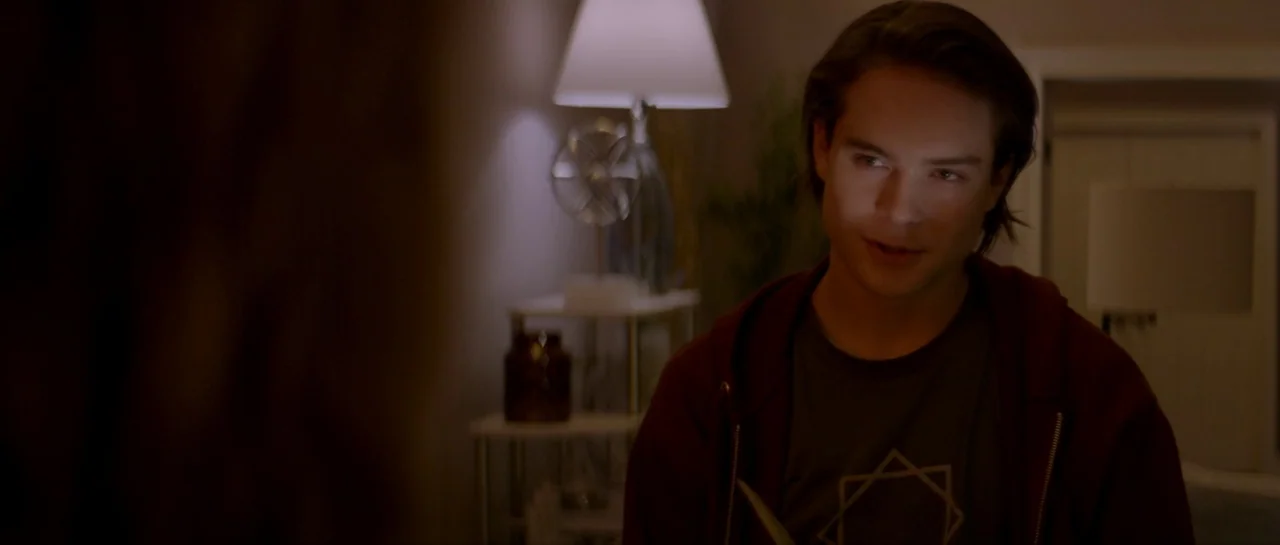 Suitable Flesh (2023) movie still: medium — An interior space features a table lamp and a blurred plant in the background. A young man in a maroon…