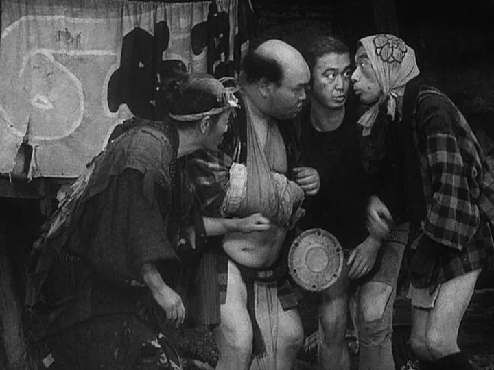 The Lower Depths (1957) movie still: medium — A dimly lit, cramped interior structure with a large fabric sign hanging overhead. Four men in…
