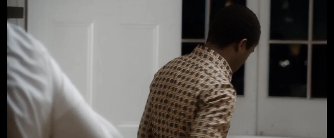 Exposed (2016) movie still: medium — An interior space features white doors and large windows looking out into darkness. A man in a gold-toned…