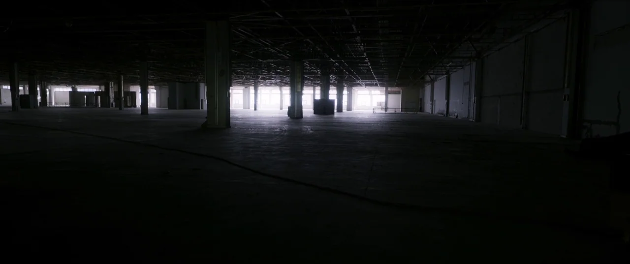 About Dry Grasses (2023) movie still: establishing — A vast, empty industrial space features concrete floors and a high, exposed truss ceiling.…