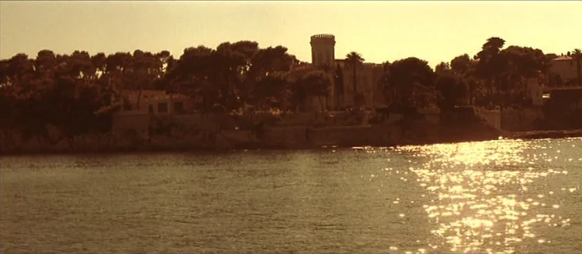 The House of Mirth (2000) movie still: establishing — A coastline features a central stone tower and dense trees overlooking the sea. Sunlight…