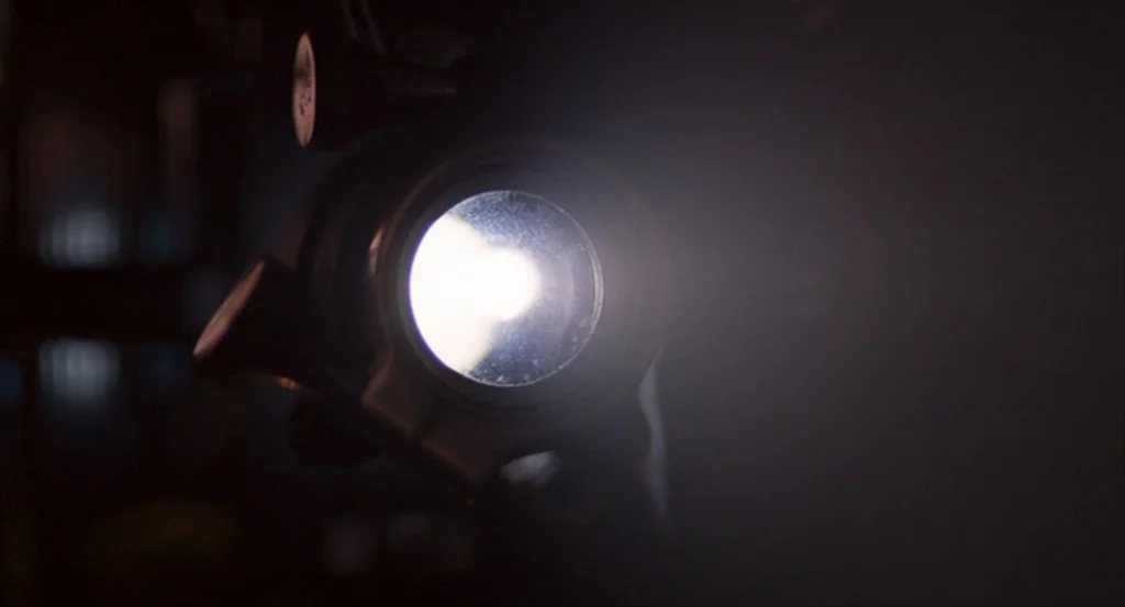The King’s Speech (2010) movie still: closeup — A close-up view of a circular lens emitting a concentrated beam of white light. The surrounding area…