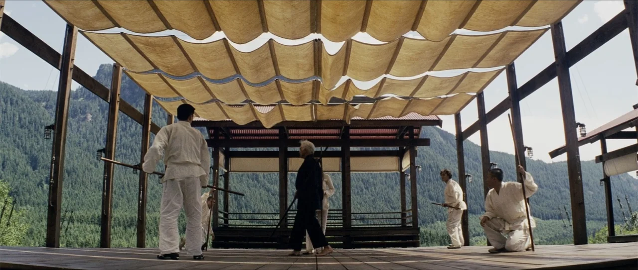 Elektra (2005) movie still: wide — An open wooden pavilion with a canvas canopy features people practicing martial arts routines. Mountain slopes…