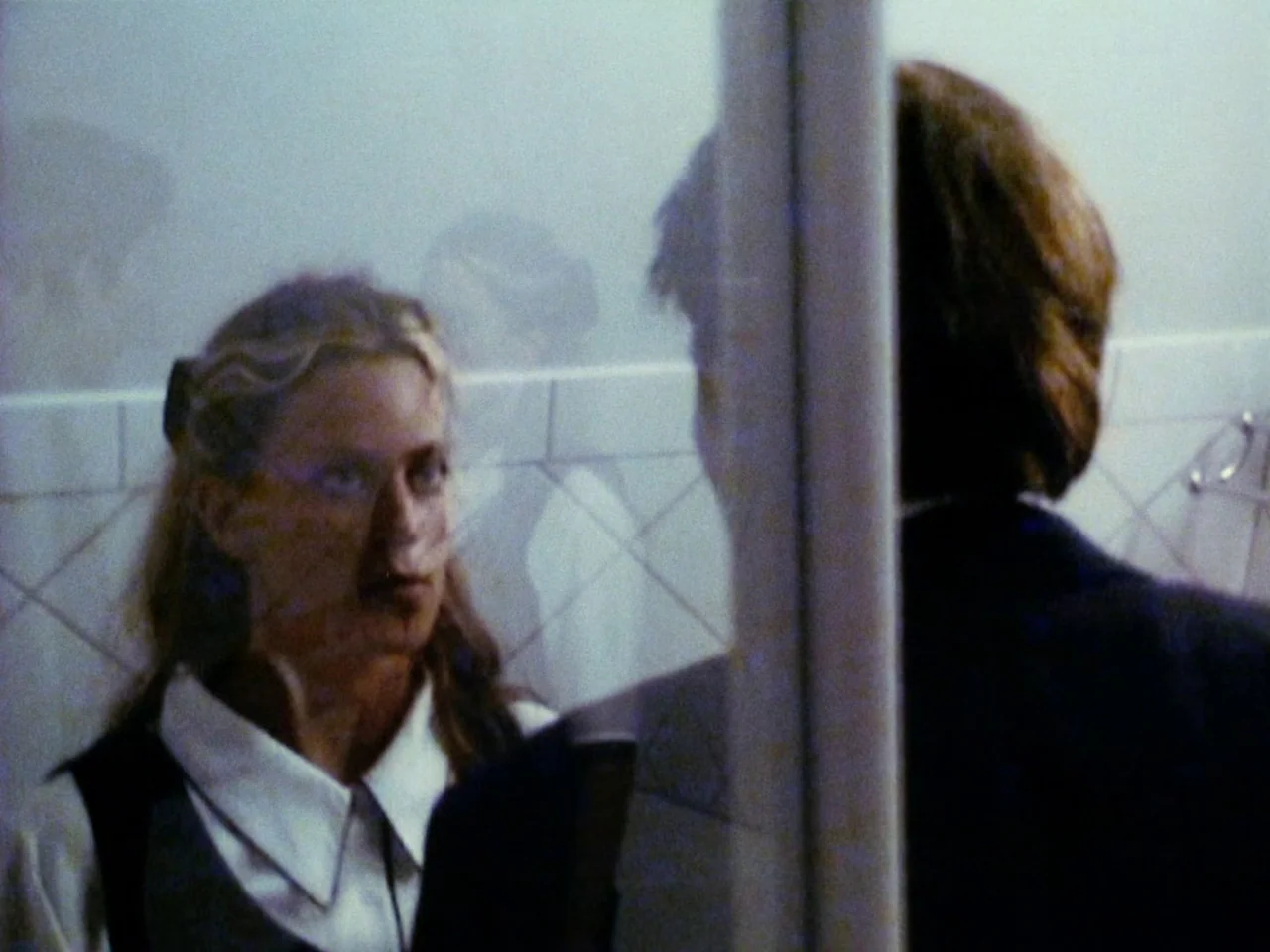 Festen (1998) movie still: medium — A tiled washroom features reflective glass surfaces and simple geometric wall patterns. A woman observes her own…