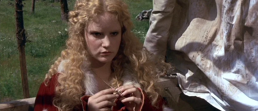 Flesh+Blood (1985) movie still: medium — A woman with blonde hair sits in a grassy outdoor area near a wagon. She holds a small object in her hands…