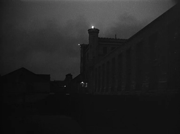 White Heat (1949) movie still: establishing — A stone-walled prison complex at night features dark, imposing silhouettes against a cloudy sky.…