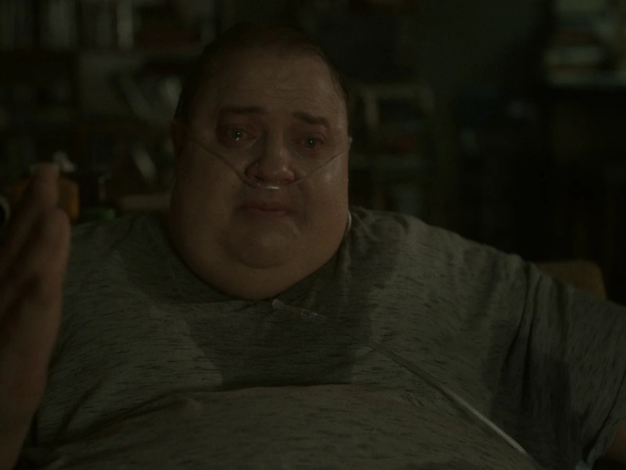 The Whale (2022) movie still: closeup — A dimly lit room with blurred bookshelves and objects in the background. A man with a nasal cannula has a…