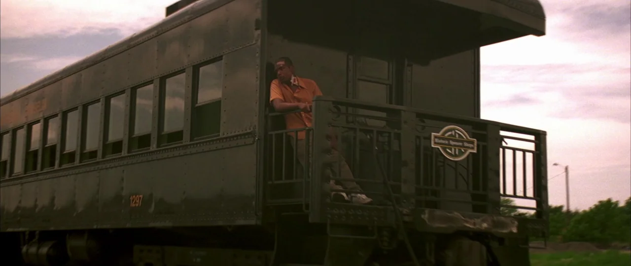 George Washington (2000) movie still: wide — A dark green vintage rail caboose sits under an overcast sky. A man stands on the rear platform leaning…