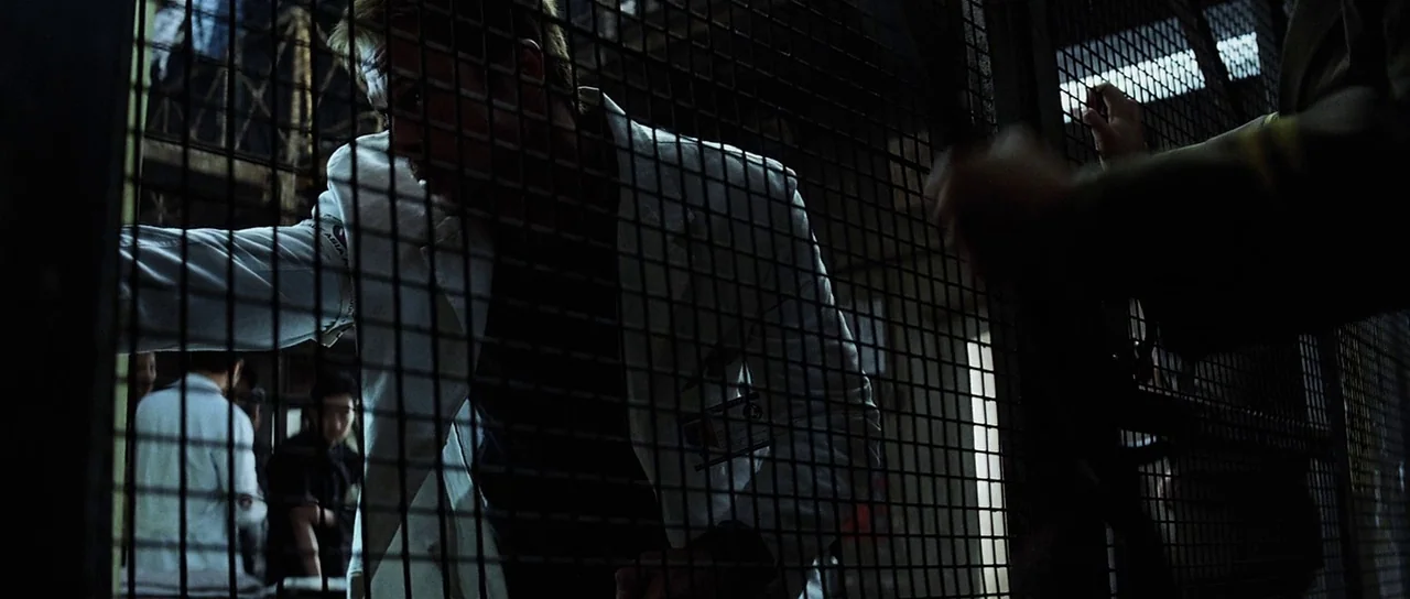 Spy Game (2001) movie still: medium — A dimly lit industrial corridor is divided by a heavy metal security mesh screen. A man wearing a white lab…