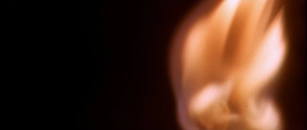 Wild At Heart (1990) movie still: closeup — A close-up view shows a single flame burning against a completely dark background. The fire flickers…
