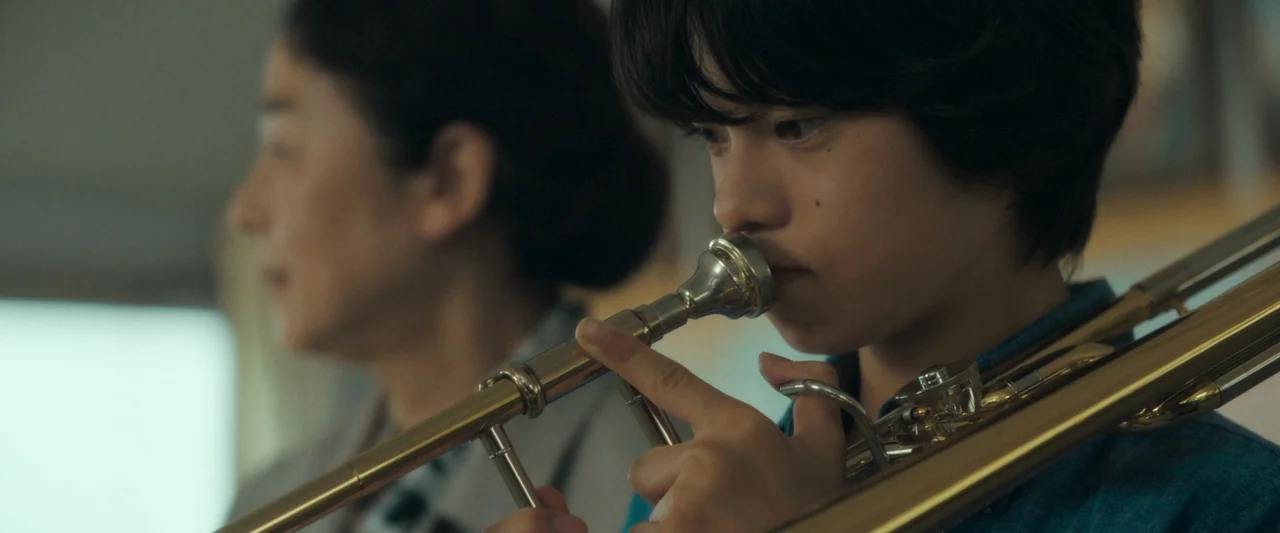 Monster (2023) movie still: closeup — Two people sit indoors with one practicing a musical instrument. A young boy in the foreground holds a brass…