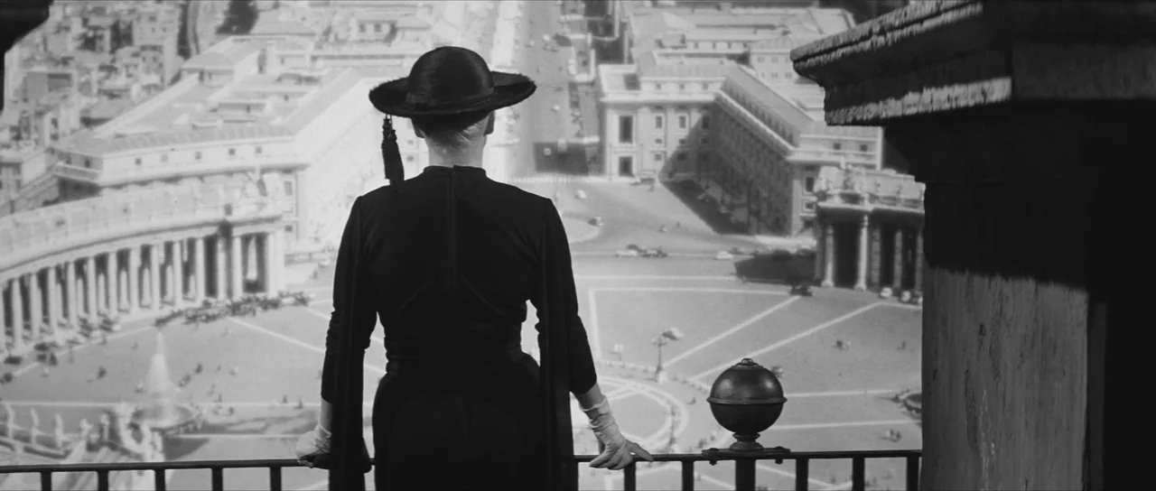La Dolce Vita (1960) movie still: fullbody — A woman stands on a stone balcony overlooking a large urban plaza. She gazes down at the sprawling…