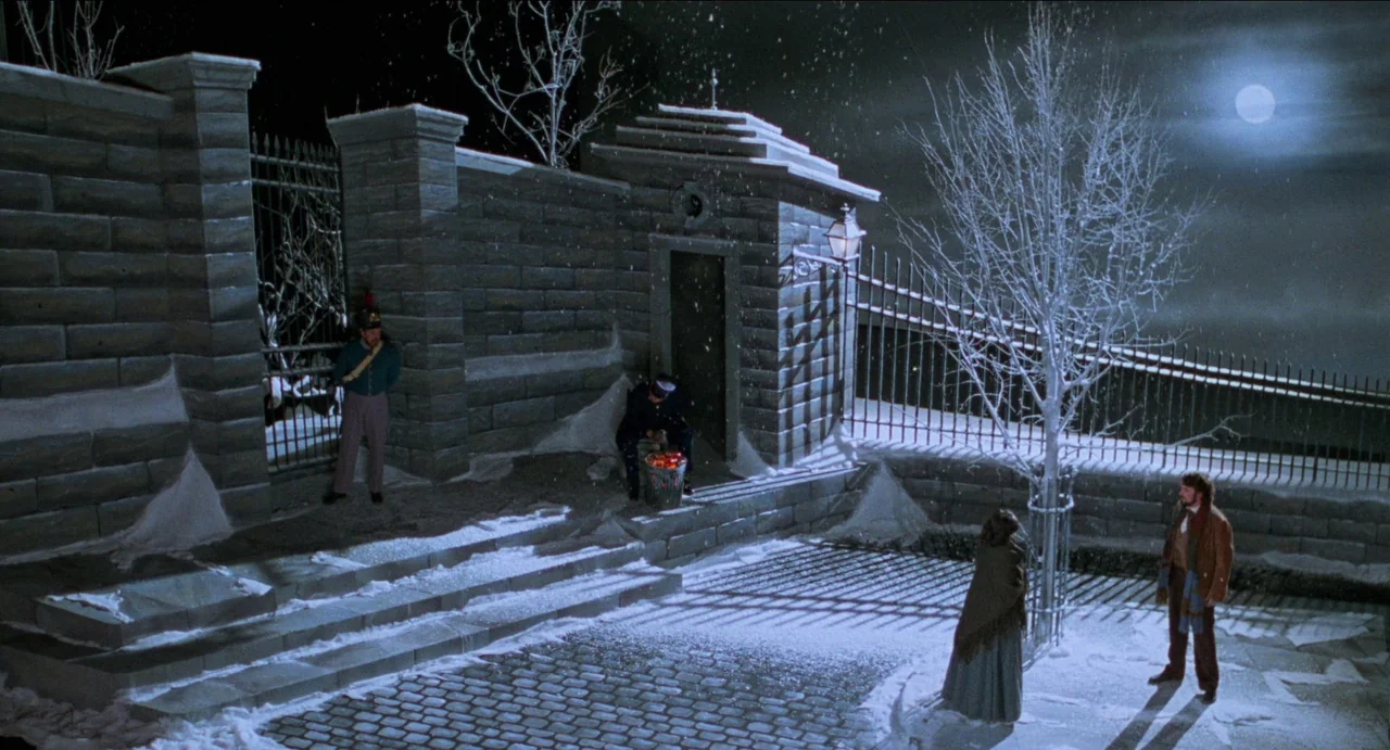 Moonstruck (1987) movie still: wide — A snowy night scene shows a stone wall with iron gates and a small guardhouse. Two people stand on the…