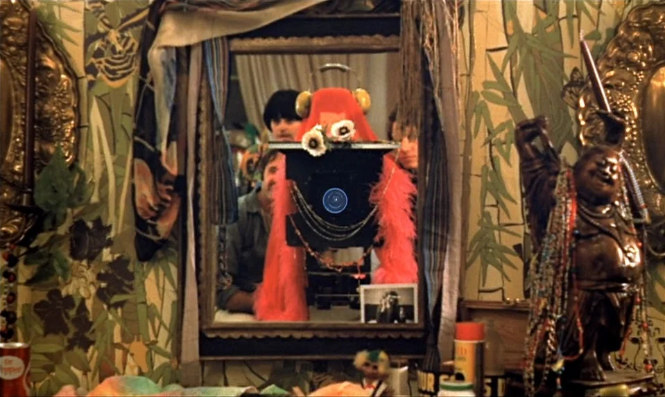Lions Love (1969) movie still: medium — A cluttered room features bamboo-patterned wallpaper and a large central mirror with draped fabrics. A man…