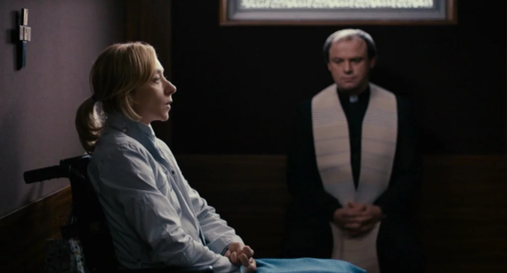 Lourdes movie still: medium — A dark, wood-paneled room with a small wooden cross mounted on the wall. A woman in a wheelchair sits opposite a…