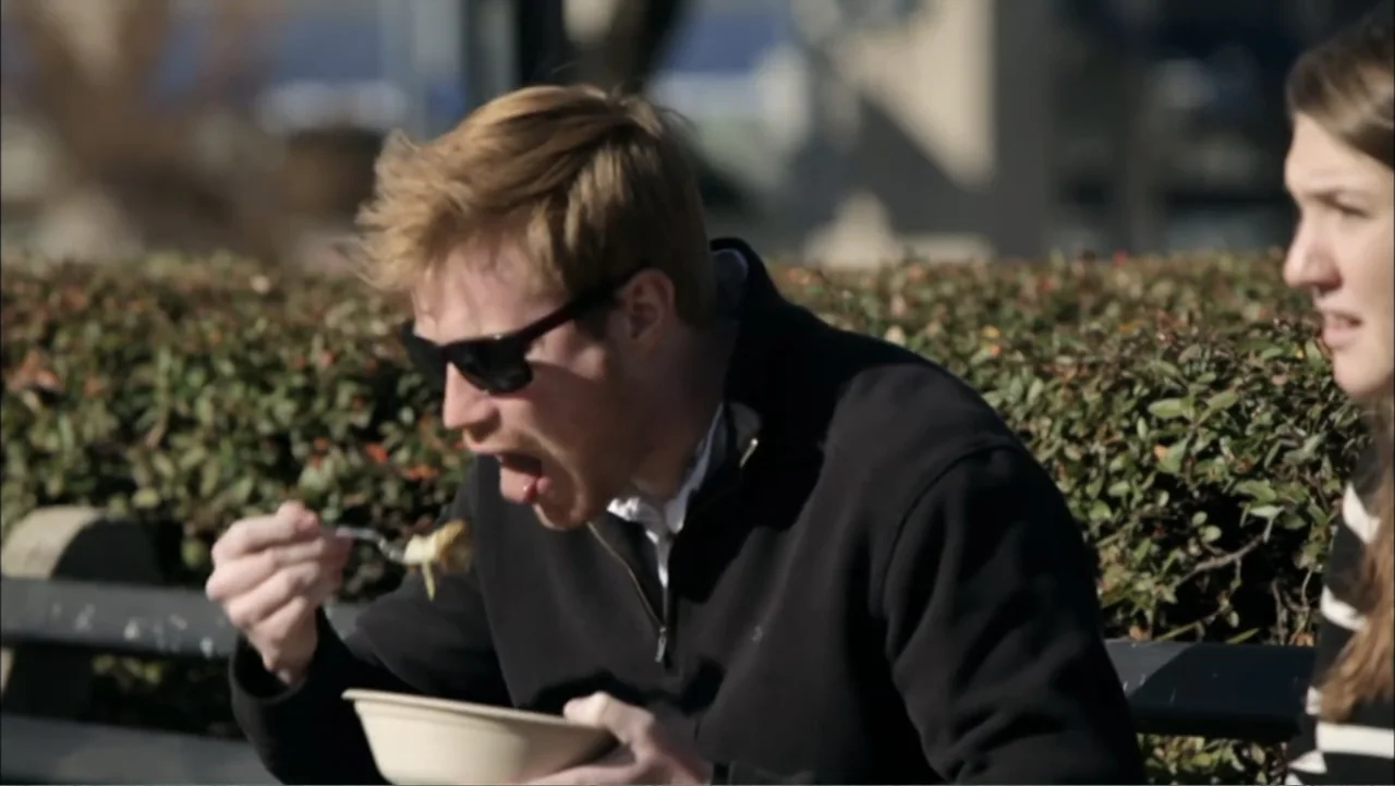 Aliens Inside Us (2013) movie still: medium — A wooden park bench sits in front of a dense green hedge on a sunny day. A man eats from a paper bowl…