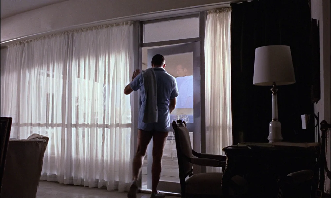 Goldfinger (1964) movie still: fullbody — A hotel room with large floor-to-ceiling windows covered by sheer white curtains. A man in blue loungewear…